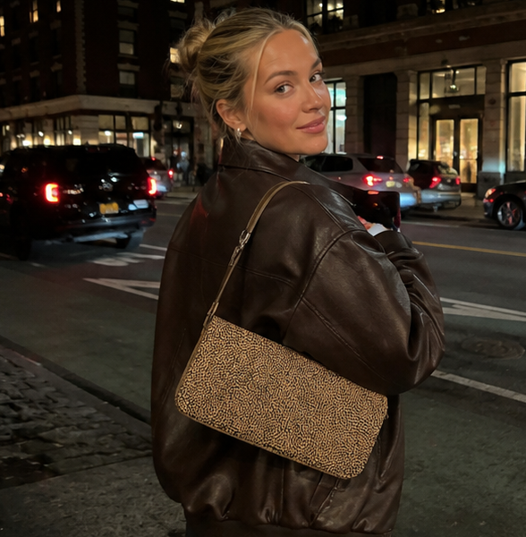 Woman in a brown leather jacket with a mustard and black NOTTEVERA handbag on a city street at night.